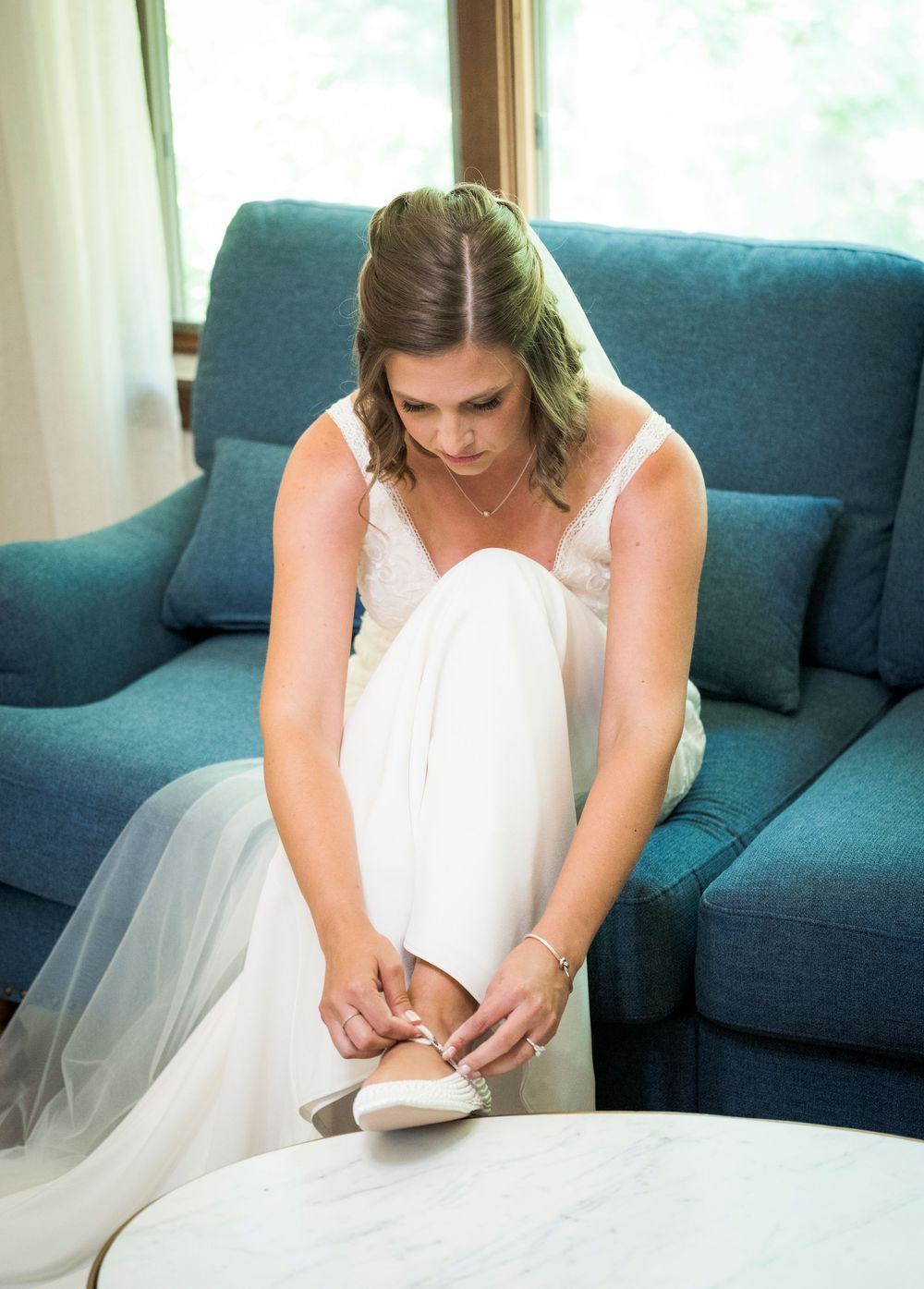 Bride in white wedding gown putting on her shoes while seated on a teal sofa before her wedding ceremony.