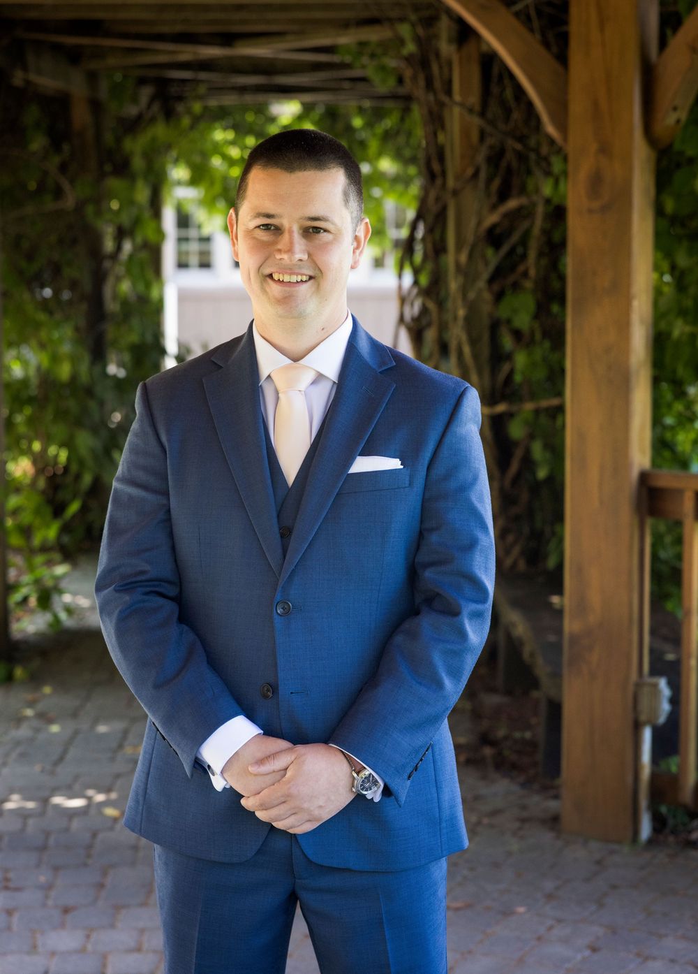 Man in blue three-piece suit with pink tie smiling under a garden pergola with lush greenery.