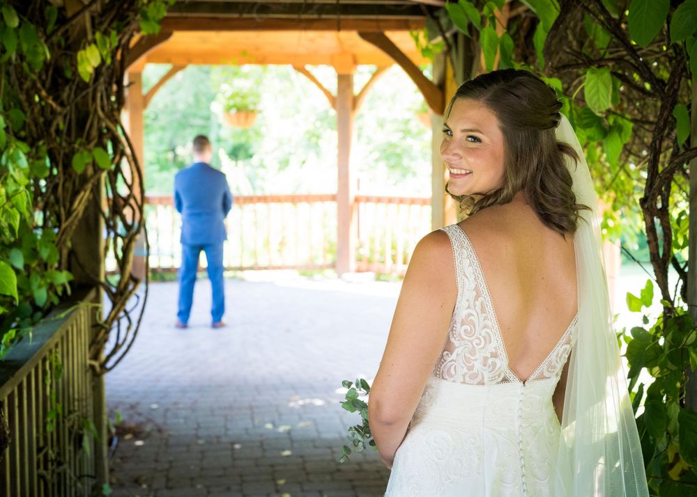 Bride in elegant backless wedding gown smiles over shoulder as groom stands under vine-covered gazebo.