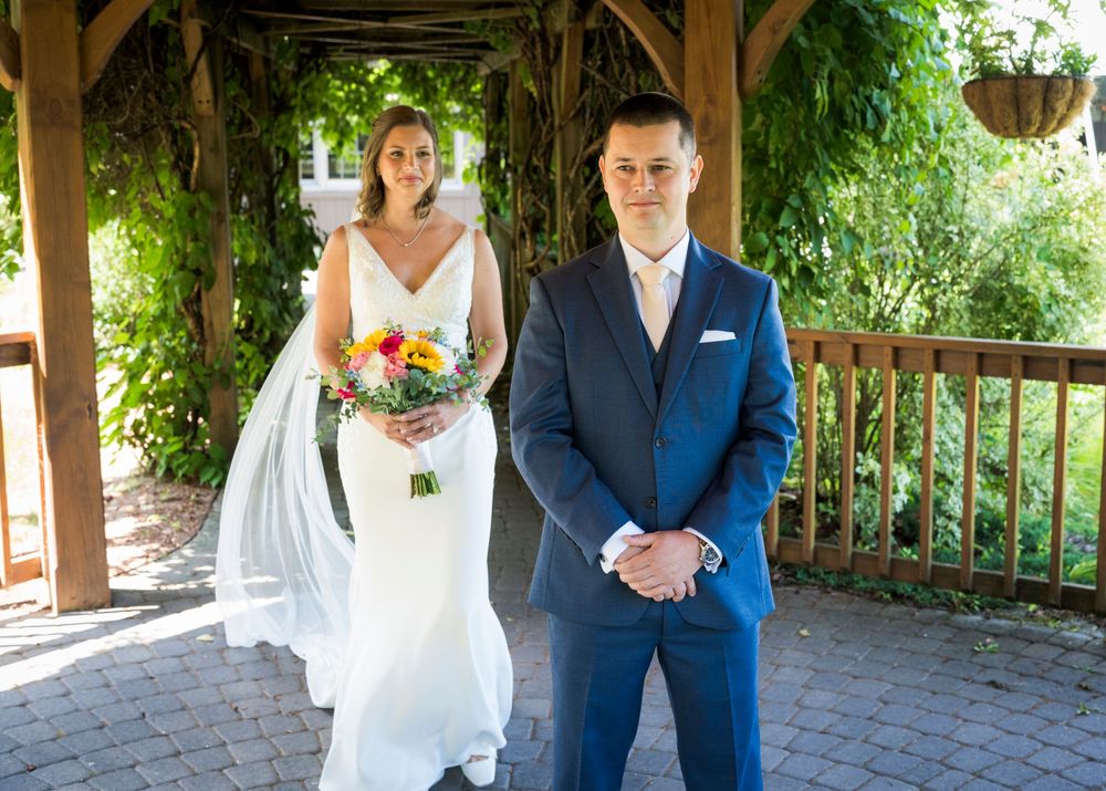 Bride in white gown holding colorful bouquet stands behind groom in blue suit under wooden pergola.