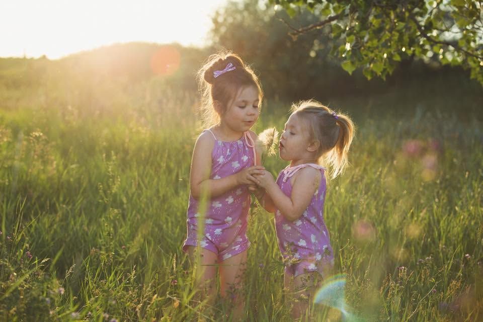 Two young girls in matching purple floral swimsuits hold hands in a sunlit green meadow during golden hour.