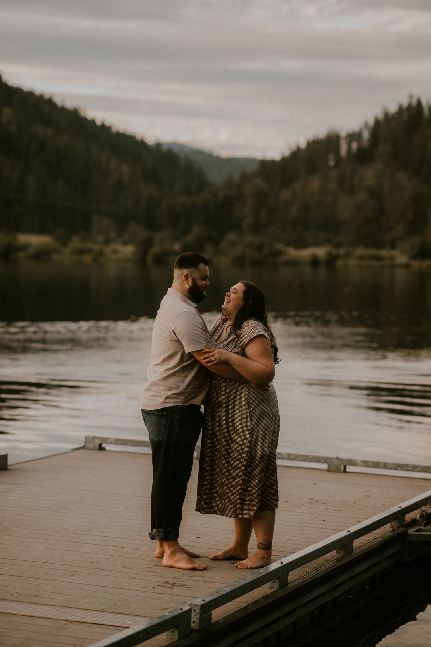 Couple embracing on a wooden dock at dusk with a serene lake and forested mountains in the background.