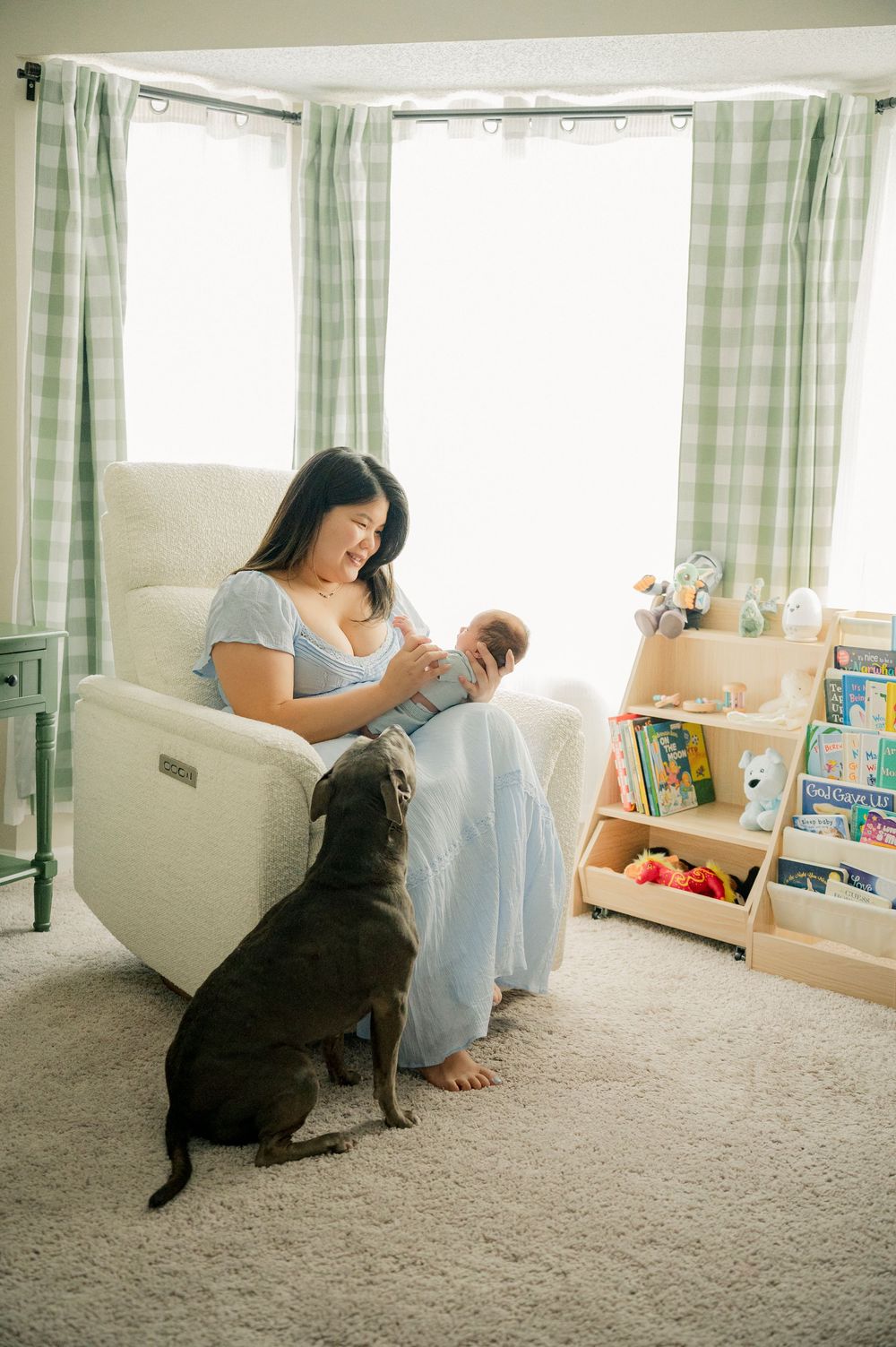 Mother holds newborn in nursery glider while dog looks on, with bookshelf and plaid curtains in background.