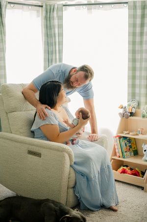 New parents admire their newborn baby in a cozy nursery, mother seated in a recliner chair wearing a blue dress.