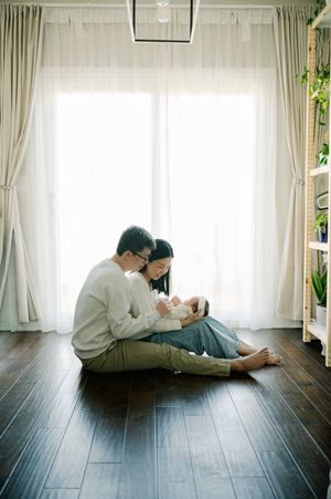 A couple sits on hardwood floor holding their newborn baby by a bright window with sheer curtains.