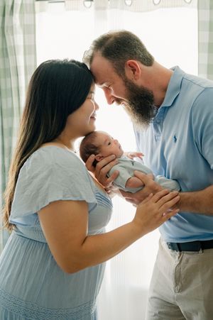 New parents lovingly hold their newborn baby together, touching foreheads in a tender moment by a sunlit window.