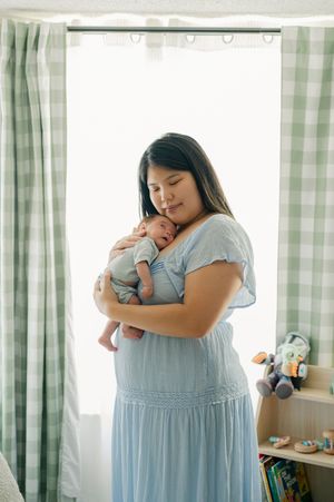 A mother in a blue dress tenderly holds her sleeping newborn baby close to her chest in a softly lit nursery.