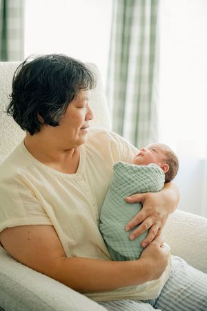 Elderly woman sitting in a white chair tenderly holding a swaddled newborn baby wrapped in a green blanket.