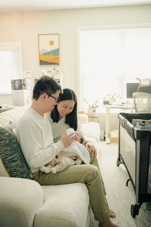 A couple sits on a couch together, lovingly gazing at their newborn baby in a bright, cozy living room.