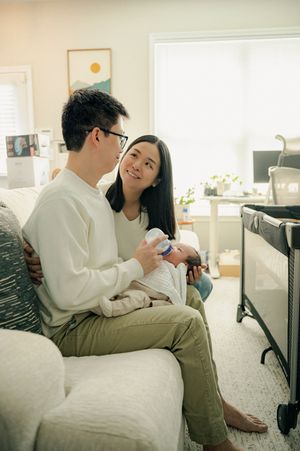 A couple sits on a couch, smiling at each other while holding their newborn baby in a bright, cozy living room.