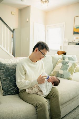 A father in glasses and cream sweater bottle-feeds his newborn baby on a cozy living room couch.
