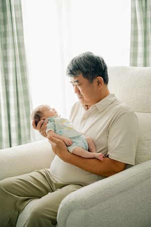 Elderly Asian man sitting in white armchair tenderly holding a sleeping newborn baby near a bright window.