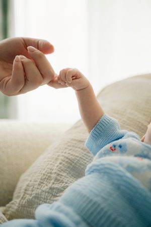 A baby in blue clothing gently grips an adult's finger on a soft white blanket.