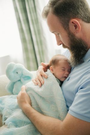 Bearded father tenderly holds sleeping newborn baby wrapped in soft blue blanket near stuffed elephant toy.