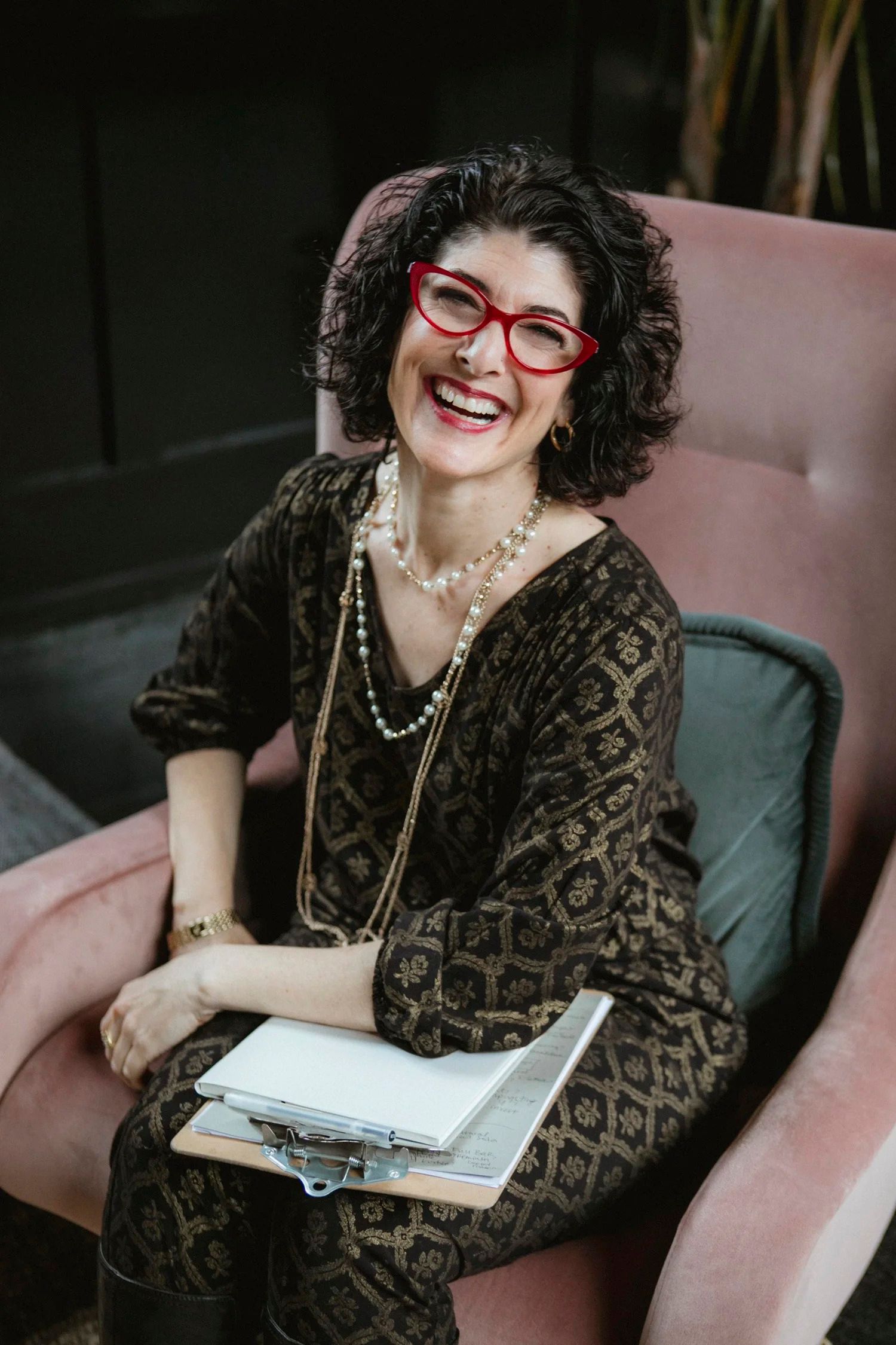 A laughing woman with red glasses and curly hair sits in a pink chair, wearing a patterned dress and pearl necklace.