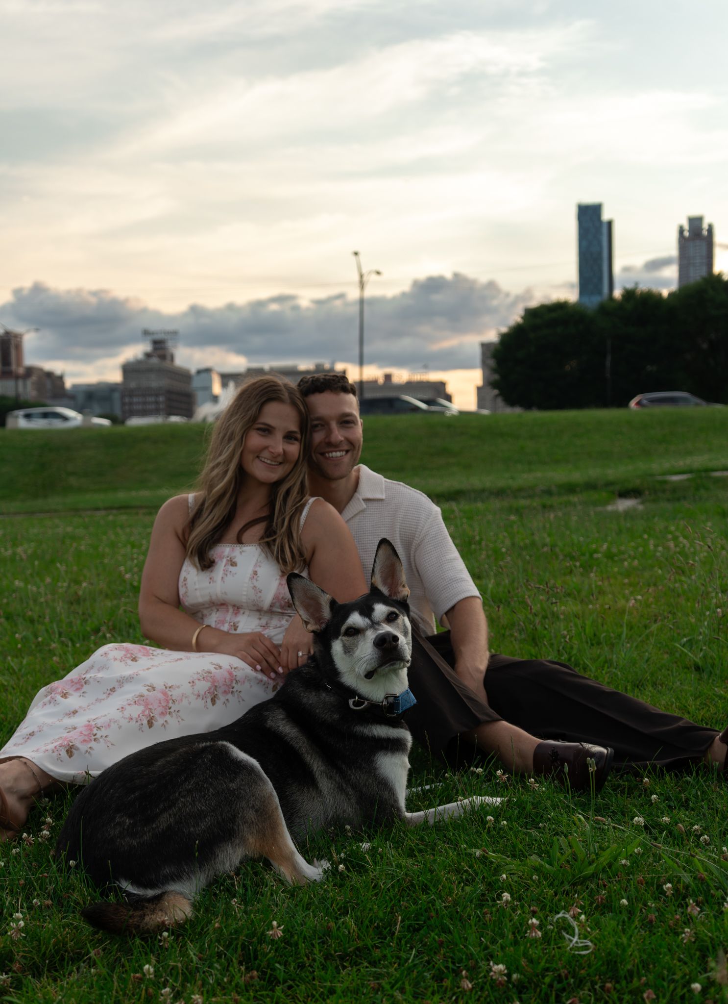 A couple sits on green grass with their Husky dog at sunset, city skyline visible in the background.