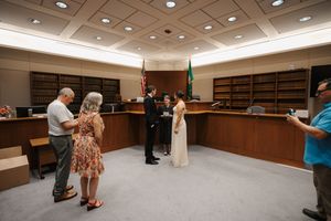 Couple exchanging vows in a courthouse ceremony with guests and officiant present, flags visible in background.