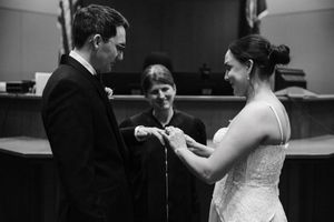 Black and white photo of bride placing wedding ring on groom's finger during ceremony with officiant.
