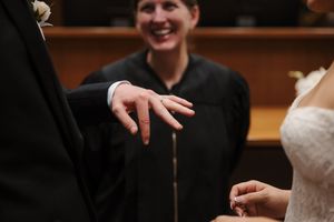 Bride placing wedding ring on groom's finger during ceremony, officiant smiling in background.