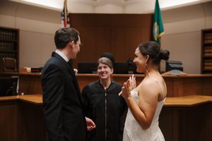 Couple exchanging vows before a judge in a courtroom wedding ceremony, bride in white dress, groom in black suit.