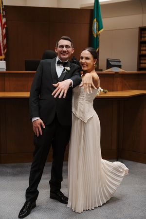 Newlywed couple in courthouse showing wedding rings, groom in tuxedo and bride in white pleated gown, Washington state flag behind them.