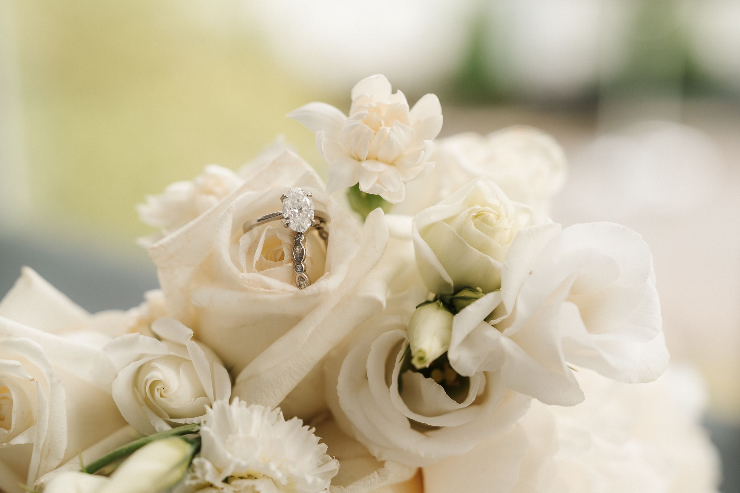 White rose wedding bouquet with diamond engagement ring and gold band nestled among ivory flowers and soft ribbon.