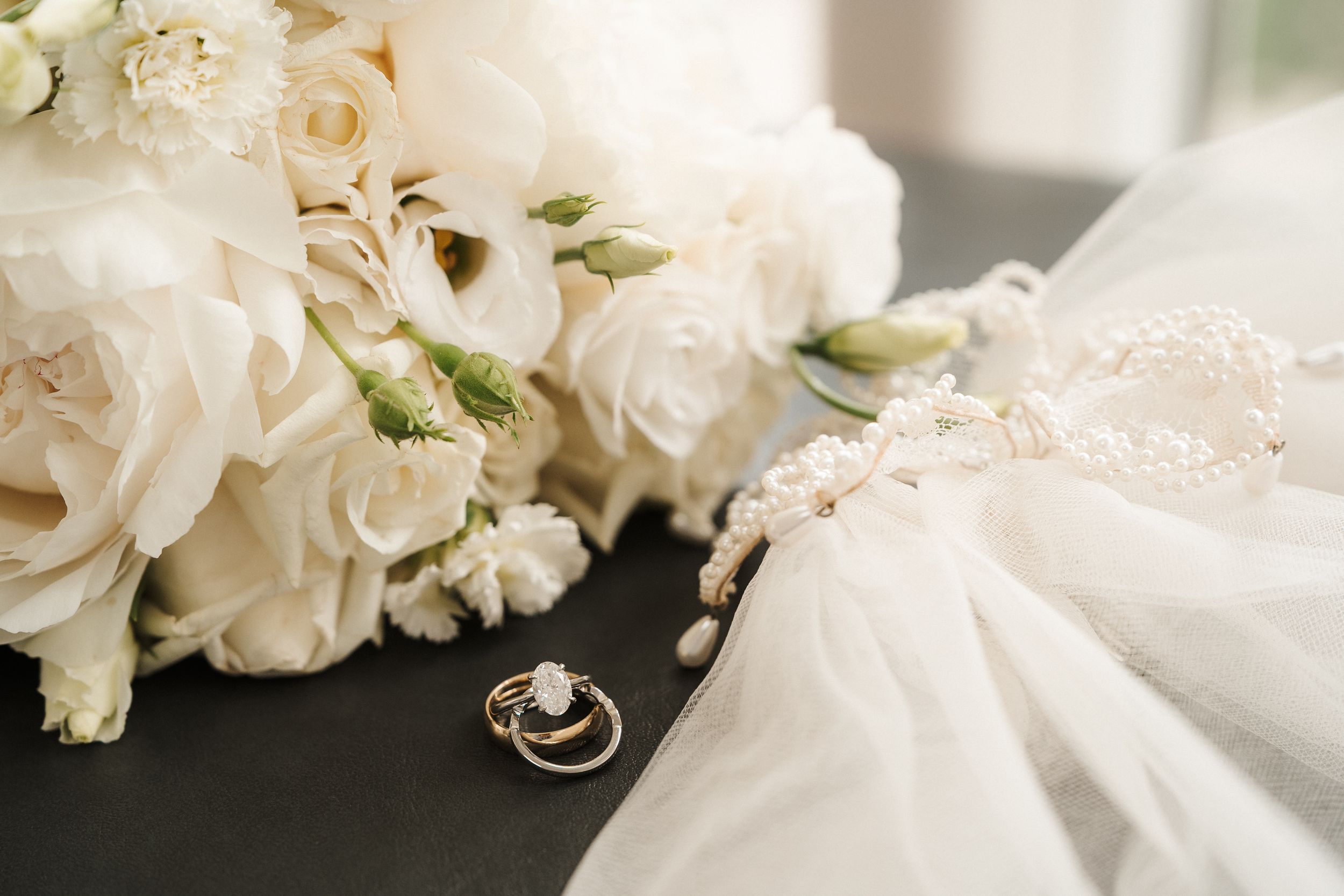 Elegant wedding flat lay with white rose bouquet, diamond engagement ring, wedding band, and pearl bridal veil on dark surface.