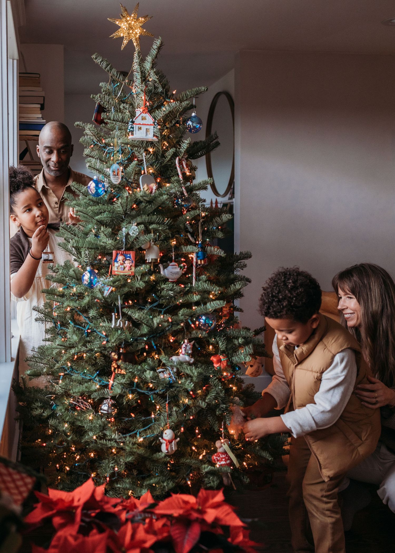 A family wearing matching red holiday sweaters sits together at a table decorating gingerbread houses for Christmas.