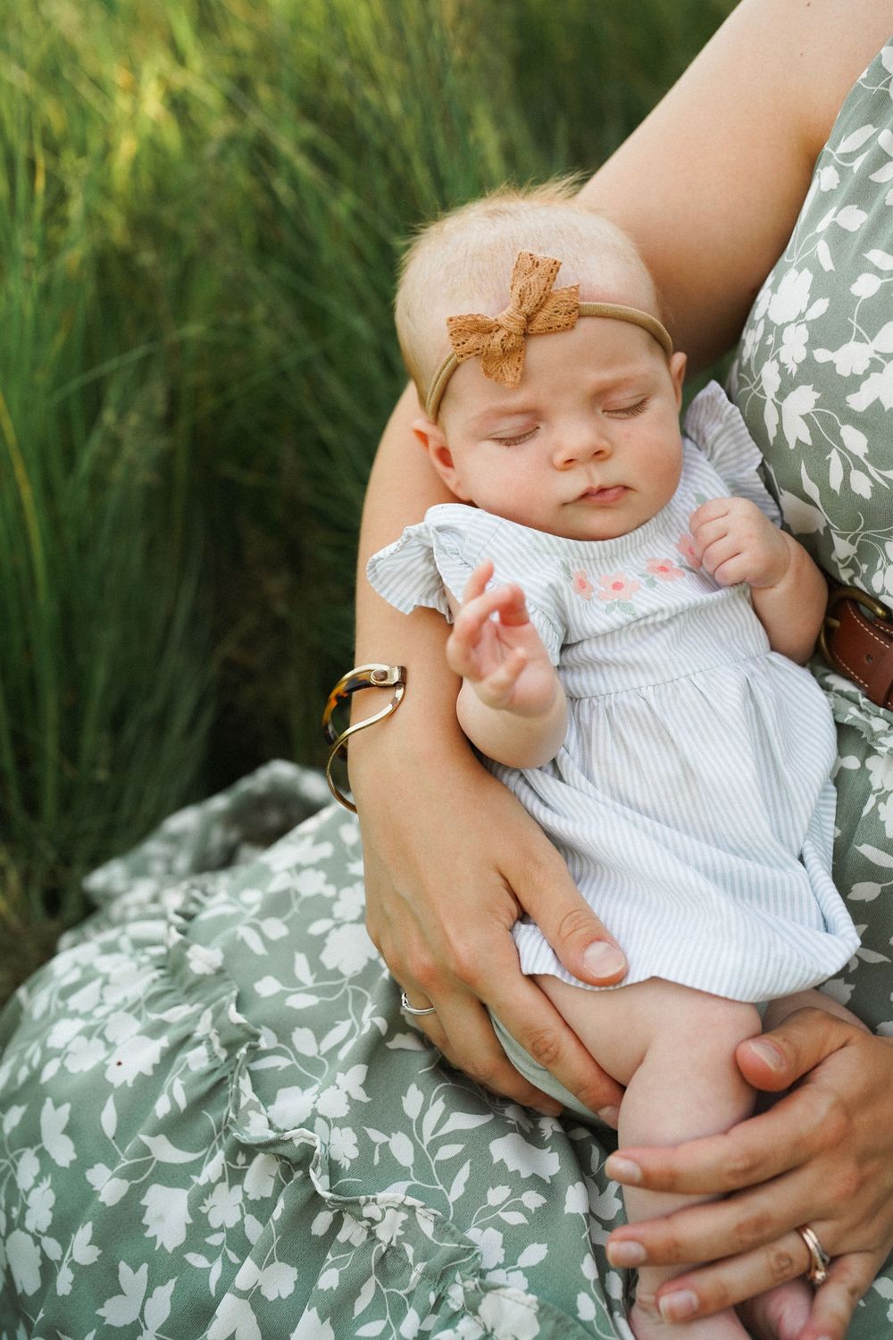 Sleeping newborn baby girl in white dress and bow headband cradled in mother's arms outdoors.