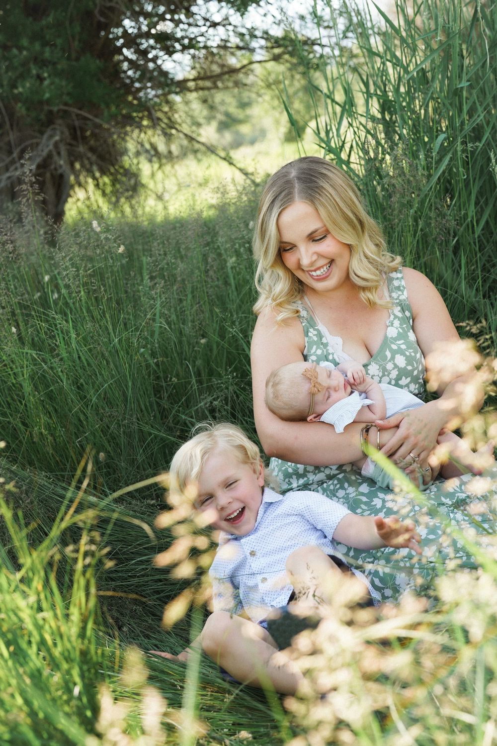 A smiling mother in a floral dress sits in tall grass, breastfeeding her newborn while her happy toddler laughs beside her.