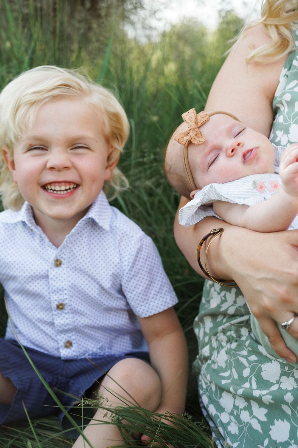 A laughing blonde toddler boy sits beside a mother holding a sleeping newborn baby girl with a bow headband outdoors.