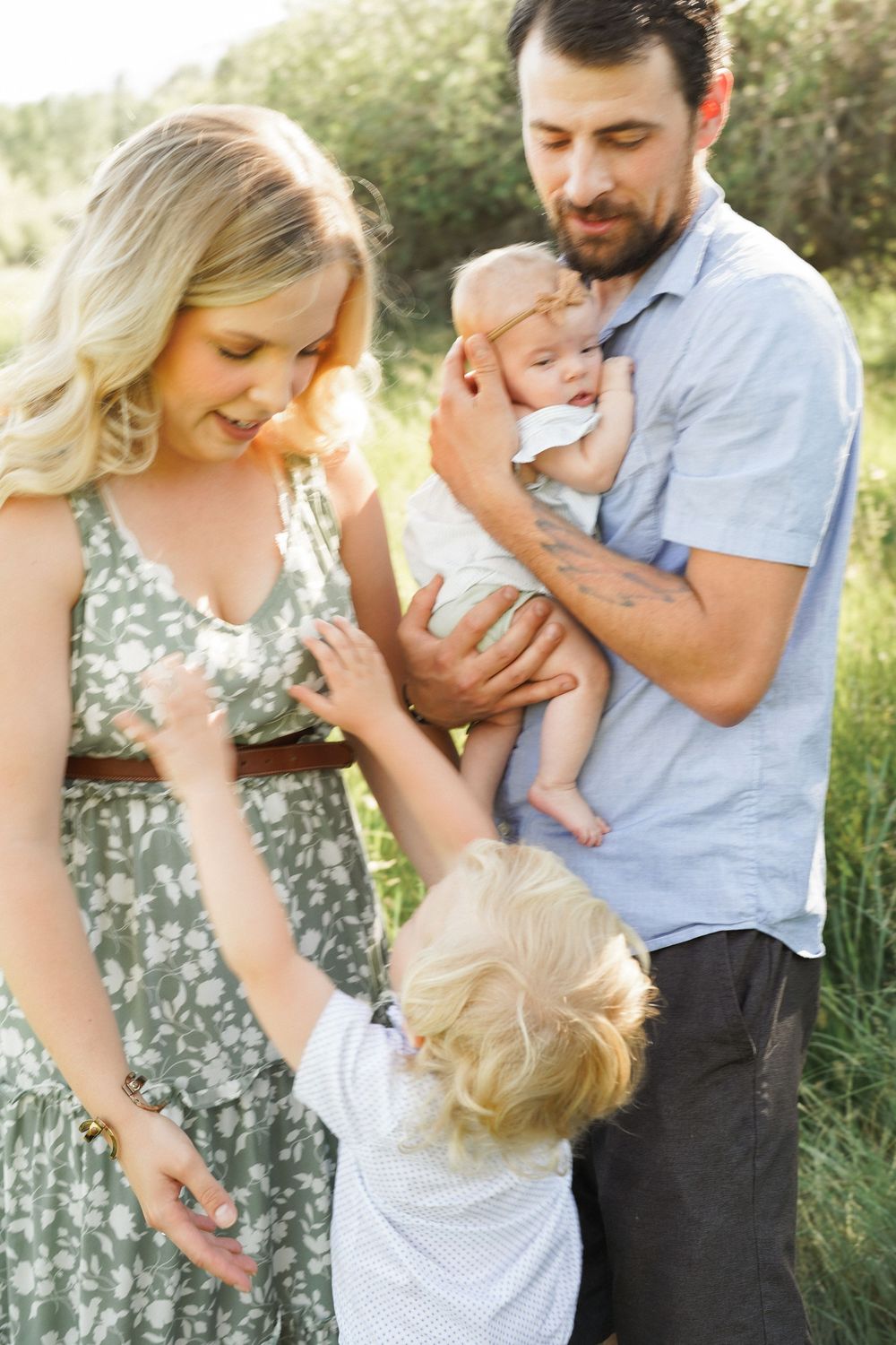 A young family with a toddler and baby enjoys a sunny outdoor moment in a green field.