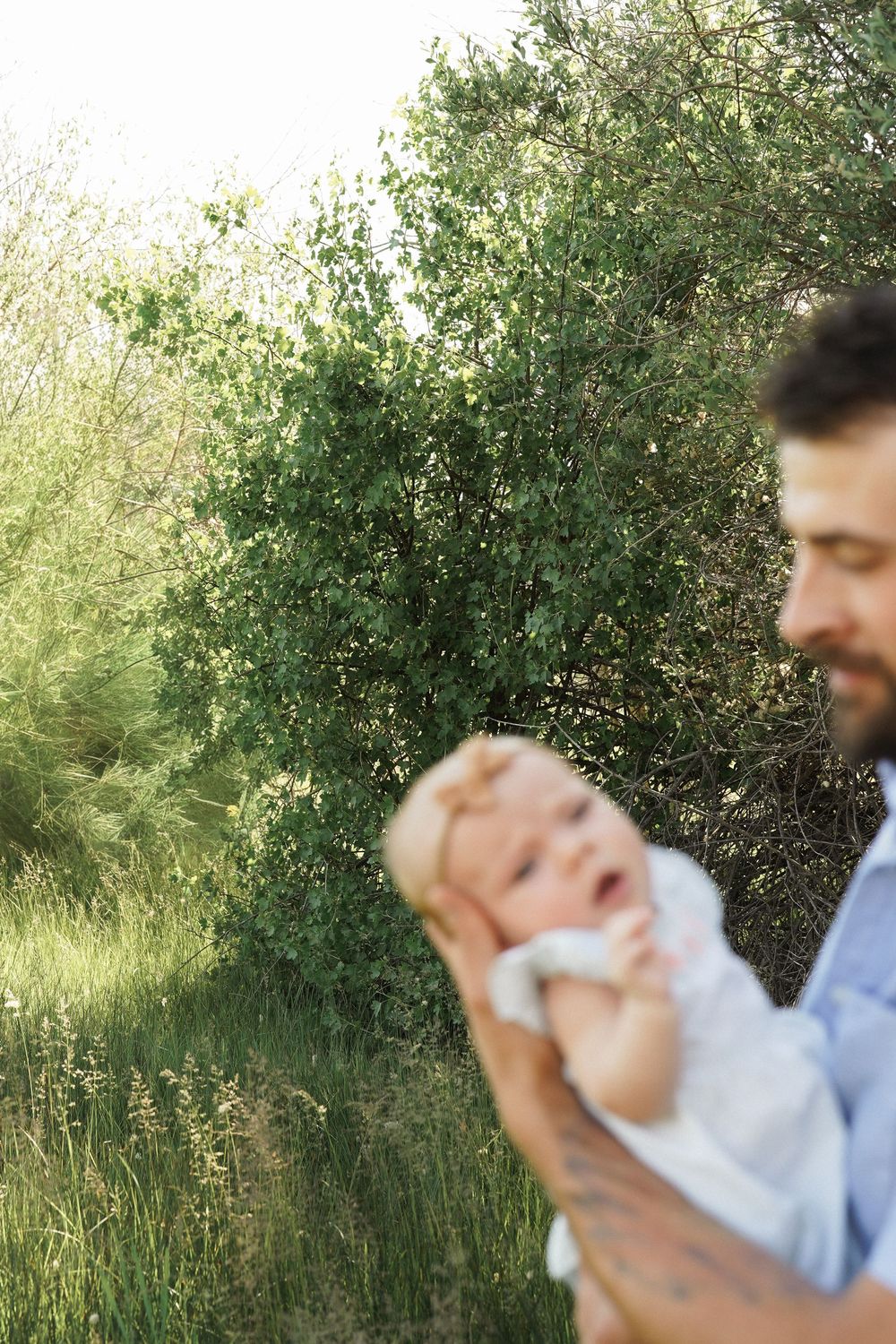 A bearded father tenderly holds his smiling baby outdoors, surrounded by lush green trees and tall grass.