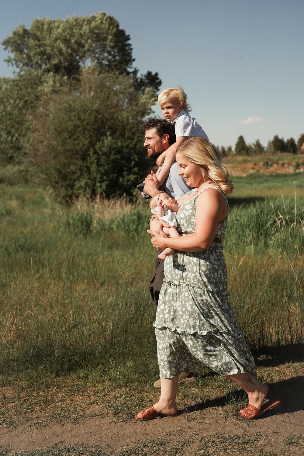Family of four walking outdoors, mother in floral dress holding newborn, father with toddler on shoulders, green meadow backdrop.