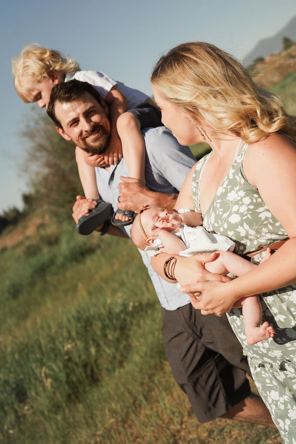 Happy family with toddler on dads shoulders and mother holding newborn baby smiling in sunny field.