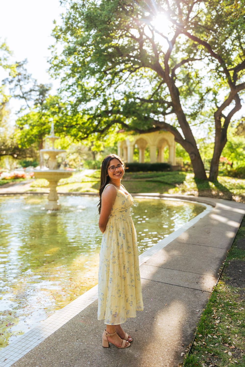 Young woman in floral yellow dress smiling by a garden fountain surrounded by lush green trees in sunlight.