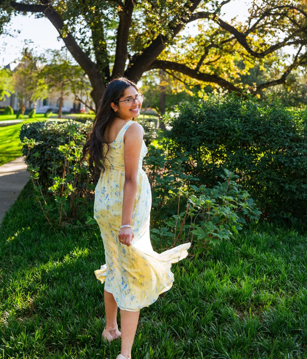 Young woman in yellow floral midi dress twirling outdoors in a lush green park on a sunny day.