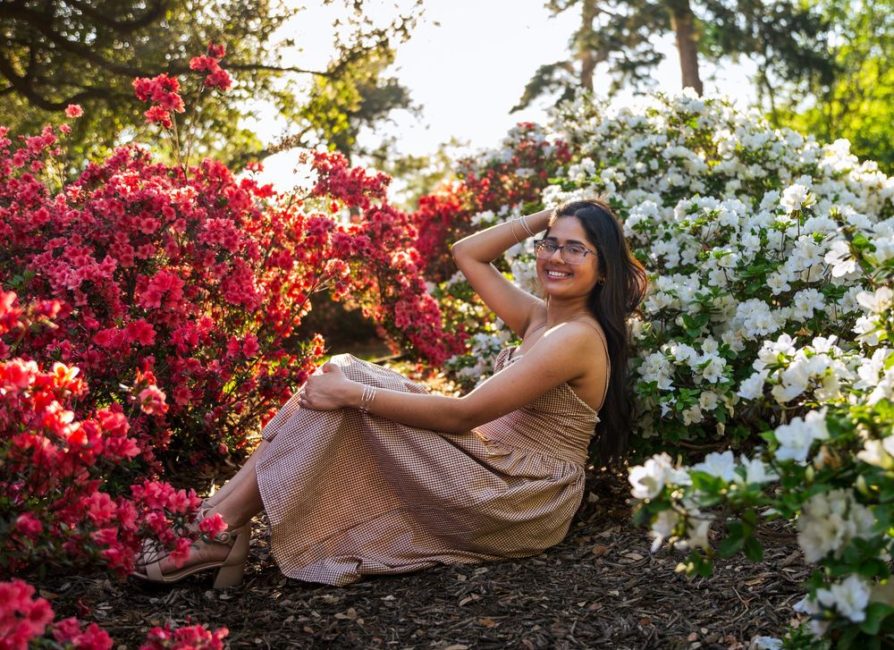 Woman in beige dress smiling among vibrant red and white azalea bushes in a lush garden setting.
