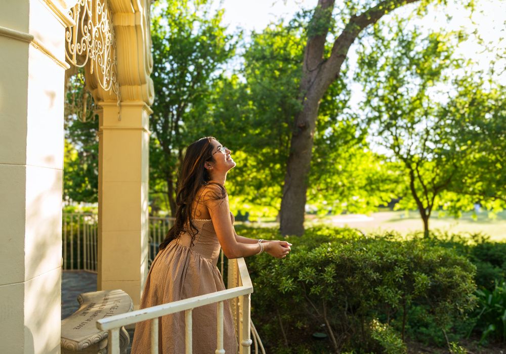 Woman in elegant gown standing on a sunlit balcony, gazing upward surrounded by lush green trees.