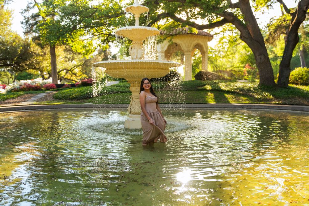 Woman in flowing dress standing in ornate fountain surrounded by lush green trees in a sunlit garden.