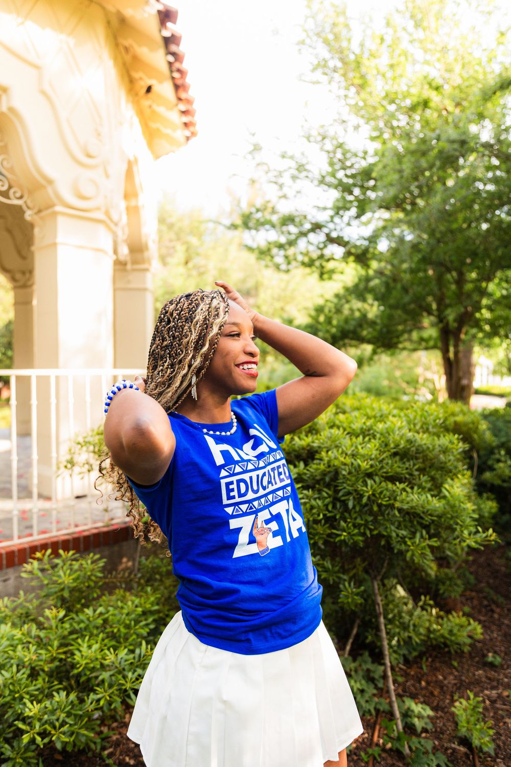 Young woman smiling in blue Zeta Phi Beta tank top and white skirt, posing outdoors near lush greenery.
