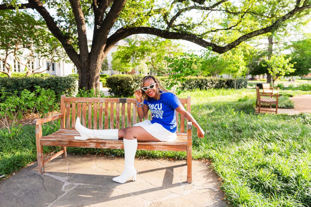 Student in blue HBCU shirt sits on wooden bench in sunny campus park with green trees and grass.
