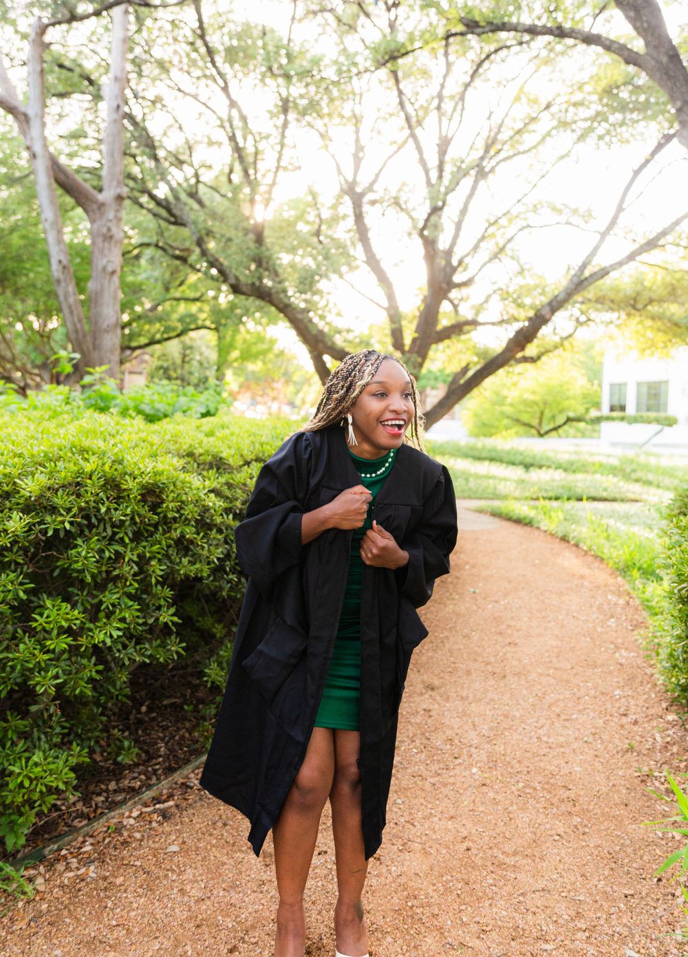 Smiling graduate in black gown and green dress walks along a garden path surrounded by lush hedges and trees.
