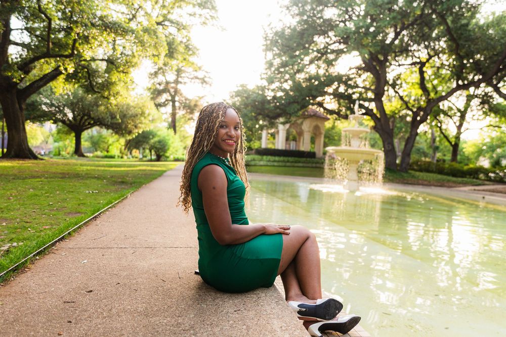 A young Black woman in a green dress sits gracefully by a fountain in a lush, sunlit park.