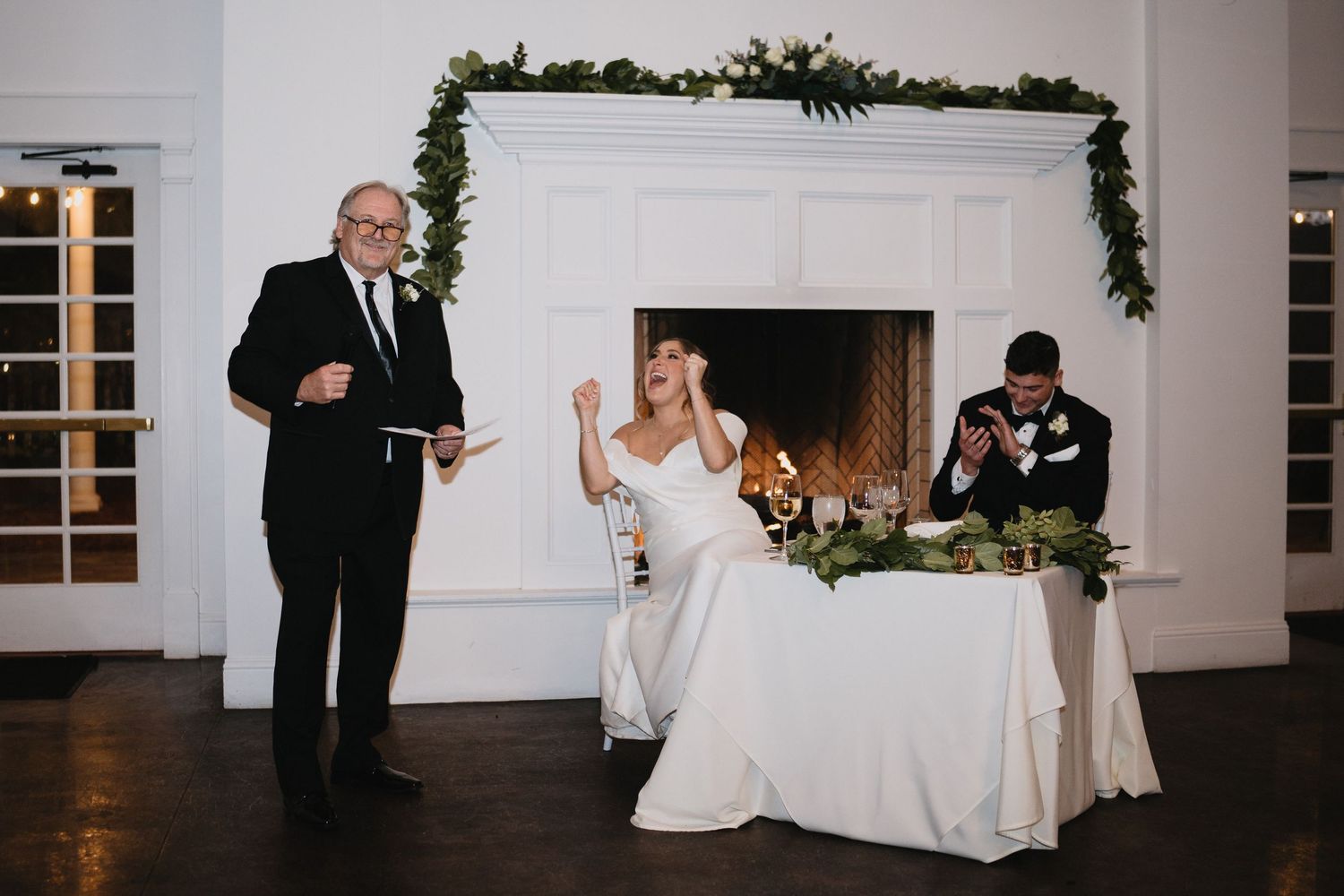 Bride and groom sit at sweetheart table laughing during wedding speech by officiant near fireplace with greenery.