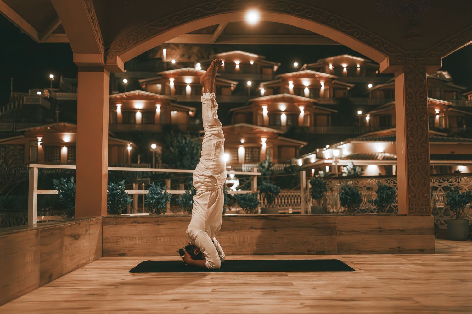 Yogi performing headstand pose on mat outdoors at night with illuminated building backdrop.