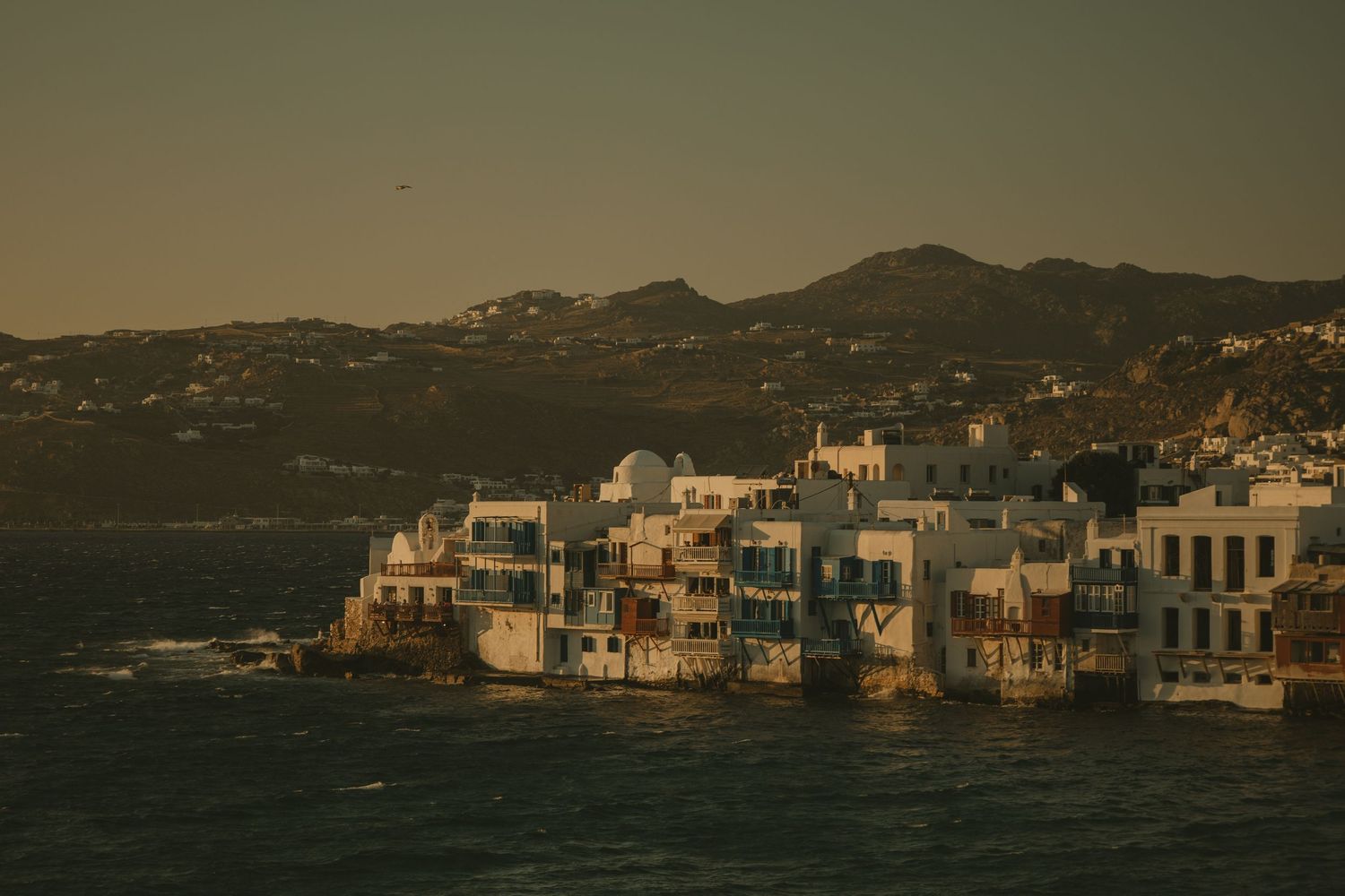 Golden hour view of Little Venice waterfront buildings in Mykonos, Greece with mountains behind.