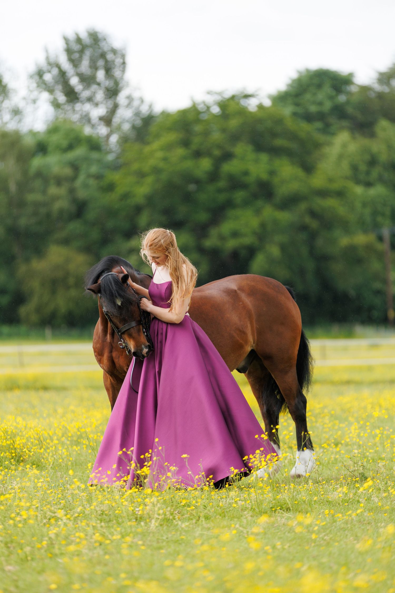 Blonde woman in jeans and boots sits on a stone wall beside a bay horse outdoors.