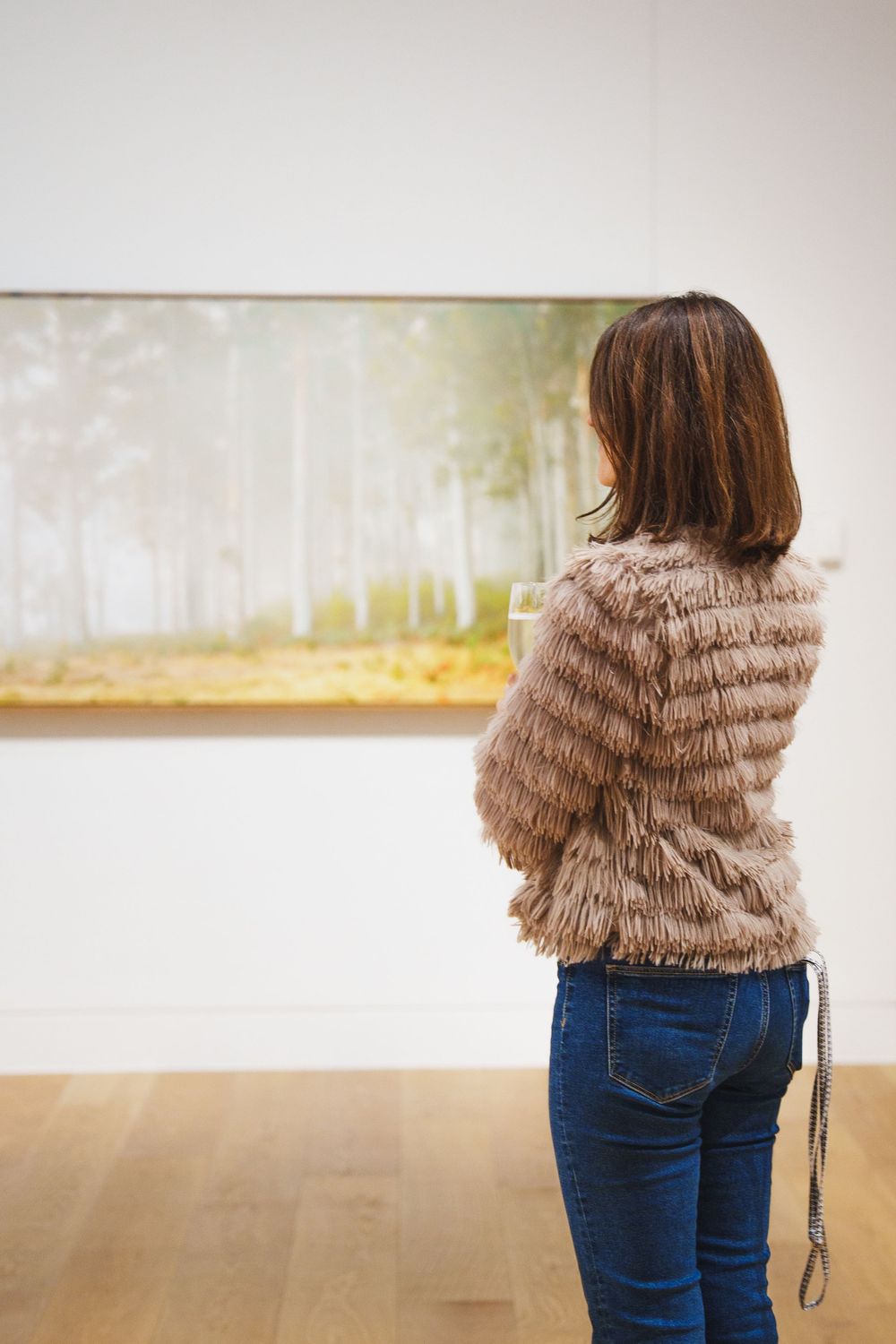Woman in fur jacket viewing misty forest landscape painting in a bright art gallery.