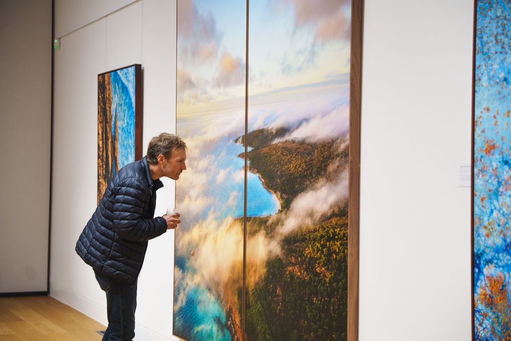 Man closely examining large aerial landscape photography triptych in a contemporary art gallery.